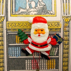 Op de afbeelding: Een vintage kerstornament met de kerstman in een rood pak en muts, met een witte baard. Hij houdt een groene kerstboom en een blauw cadeau vast. Het ornament heeft zwarte laarzen en een rood touwtje om op te hangen.