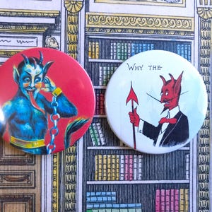 Op de afbeelding: Twee ronde buttons met duivelillustraties. Eén button toont een blauwe duivel met een lange tong op een rode achtergrond. De andere heeft een rode duivel in een pak die een speer vasthoudt, met de tekst "WHY THE" op een witte achtergrond.