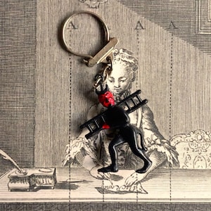 Könnte beinhalten: Ein Metallschlüsselanhänger mit einer kleinen, schwarz gekleideten Figur mit rotem Gesicht und Hörnern, die eine schwarze Leiter auf den Schultern trägt. Der Anhänger ist mit einer kurzen Kette und einem dreieckigen Verbindungsstück an einem silbernen Ring befestigt.