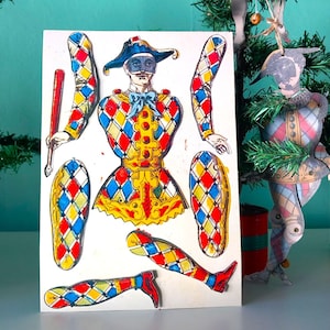 Puede incluir: Una muñeca de papel vintage de un arlequín con un disfraz de rombos, con brazos, piernas y un bastón separados. El arlequín lleva un sombrero y una máscara azules. Un adorno de arlequín similar cuelga en el fondo, decorado con vegetación artificial.