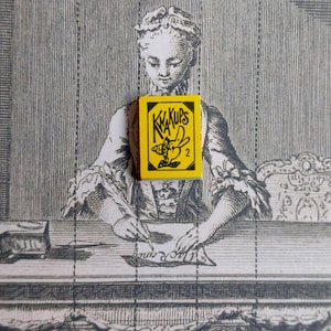 Op de afbeelding: Een gele en zwarte KWA KUPS lucifersdoos is gecentreerd boven een vintage gravure van een vrouw die aan een bureau schrijft. De lucifersdoos heeft een cartoonbij en het nummer 2. De achtergrond is een gedetailleerde zwart-wit illustratie.