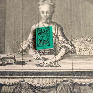 Könnte beinhalten: Ein kleines, türkisfarbenes Notizbuch mit dem schwarzen Schriftzug "Silly Dillies". Das Notizbuch befindet sich vor einer Vintage-Radierung einer Person, die an einem Schreibtisch sitzt. Das Bild hat eine Vintage-Ästhetik.