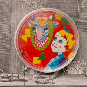May include: A round plastic puzzle game featuring a red background with a green-faced creature and a blonde child. The game includes small metal balls that move through holes in the illustrations. The text "MADE IN HONG KONG" is printed on the surface.