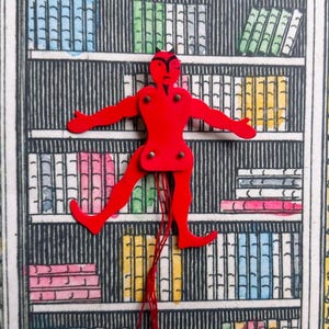 以下が含まれることがあります： 悪魔のような外観の赤い関節人形が、本棚のイラストを背景に配置されています。人形は腕と脚を広げ、関節と赤い糸が見えます。背景には、カラフルな本でいっぱいの本棚の詳細な図面が描かれています。