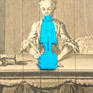 Könnte beinhalten: Eine hellblaue Kunststofffigur einer stehenden Gestalt mit Umhang, die einen Dreizack hält und auf einer Basis aus stilisierten Flammen steht. Die Figur ist vertikal ausgerichtet und weist eine glatte, geformte Oberflächenstruktur auf.