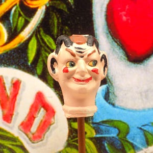 Könnte beinhalten: Eine skurrile, handbemalte Keramikfigur eines Clowns mit einem schelmischen Grinsen. Der Clown hat eine rote Nase, bemalte Augenbrauen und einen roten Mund. Die Figur ist auf einem Holzstab montiert.
