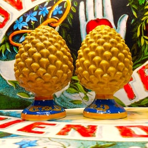 May include: Two ceramic pinecone-shaped ornaments featuring a textured yellow upper section and a blue base decorated with gold floral patterns and white dots. The objects are symmetrical, standing upright with a rounded top and a flared pedestal.
