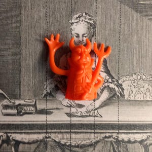 May include: A bright orange plastic figurine of a devil character with horns, a raised right hand, and a trident held in the left hand. The figure has a moulded face with a pointed chin and a textured body, presented as a small, solid toy object.