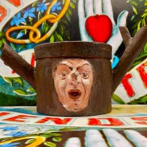 Op de afbeelding: Een bruine, cilindrische container met een gebeeldhouwd gezicht en takachtige handvatten. Het gezicht heeft een verbaasde uitdrukking. De container staat tegen een kleurrijke achtergrond met het woord "LOVE" zichtbaar.