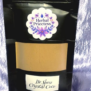 May include: A black pouch containing a tan powder, visible through a clear section. The pouch features a white label with "He Shou Crystal Coco" and a floral sticker reading "Herbal Priestess". The background is a light purple fabric.