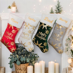 May include: Four Christmas stockings in red, white, green, and gray, each with a white faux fur cuff. Gold snowflakes and "Merry Christmas" are printed on the body. Names "Mom", "Sophia", "Dad", and "Henry" are embroidered in gold.