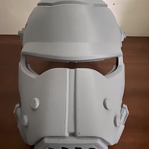 May include: A gray, 3D-printed helmet with a visor and ventilation slots. The helmet has a rounded top with circular details and several protruding circular accents. The face mask has a central vertical ridge and curved side details.