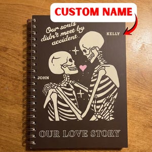 Op de afbeelding: Een spiraalgebonden notitieboek met een donkerbruine kaft met een illustratie van een skeletpaar. De tekst "Our souls didn't meet by accident" en "Our Love Story" staat op de kaft. De namen "John" en "Kelly" zijn ook zichtbaar.