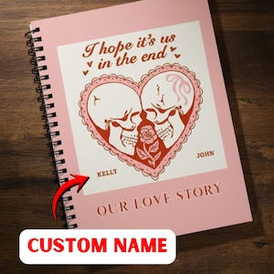 Op de afbeelding: Een roze spiraalgebonden notitieboek met een decoratief hartontwerp met twee schedels en een roos. De tekst "I hope it's us in the end" staat boven het hart en "Our Love Story" eronder. Het notitieboek heeft een "Custom Name" label.