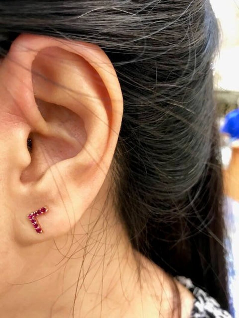May include: A close-up of a person's ear with a small red ruby letter 'L' earring.