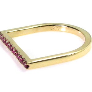 Op de afbeelding: Een gouden ring met een rechthoekige band en een rij kleine rode edelstenen. De ring is aan &eacute;&eacute;n kant open, waardoor hij aan verschillende vingers gedragen kan worden.