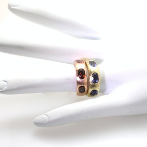 May include: Two gold rings with gemstones. The top ring has five blue gemstones and the bottom ring has five red gemstones.