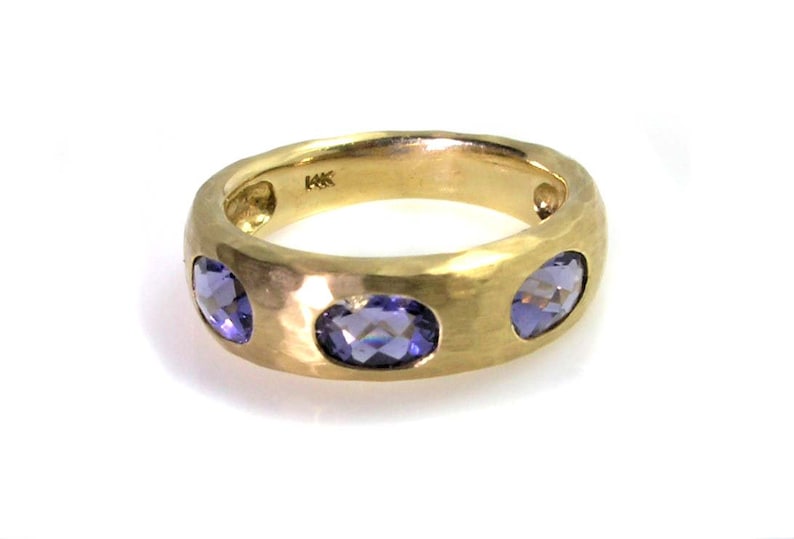 May include: A gold band ring with three purple gemstones set into the band. The band has a hammered texture.