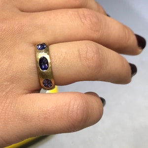 May include: A gold band ring with three oval-shaped purple gemstones set into the band.