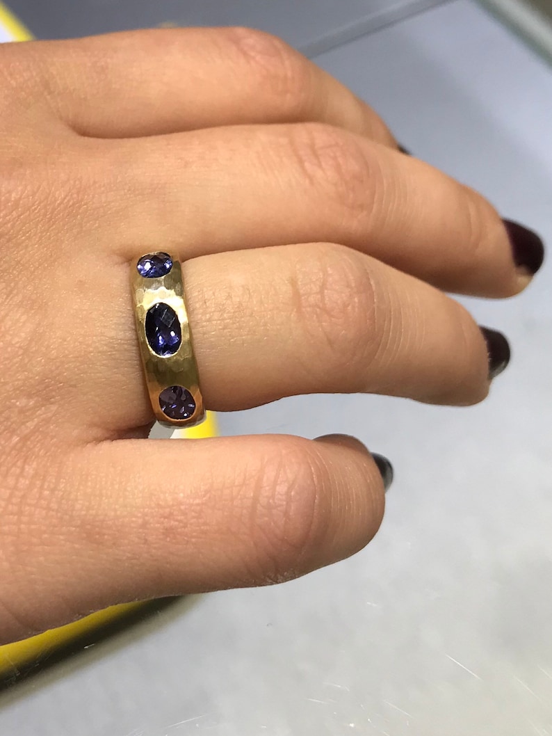 May include: A gold band ring with three oval-shaped purple gemstones set into the band.