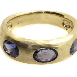 May include: A gold band ring with three oval-shaped purple gemstones set into the band.