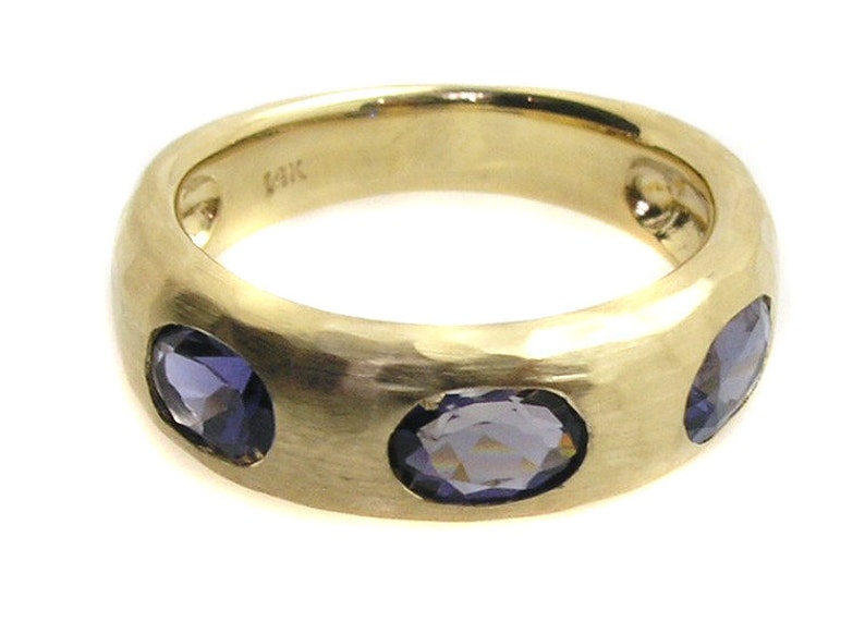 May include: A gold band ring with three oval-shaped purple gemstones set into the band.