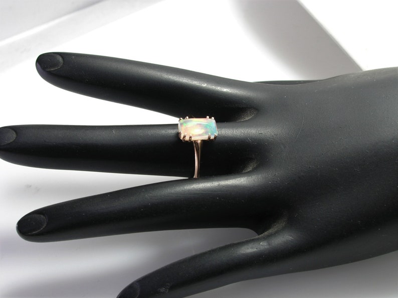 May include: A gold ring with a rectangular opal gemstone. The ring is set on a black ring display stand.
