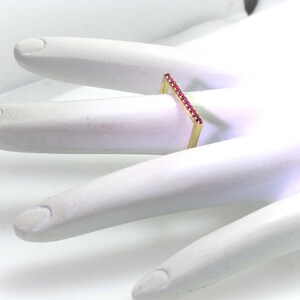 Op de afbeelding: Een gouden ring met een rechthoekige band bezet met kleine rode edelstenen.