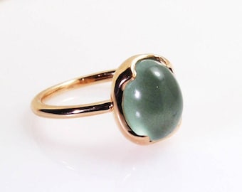 May include: A rose gold ring featuring a large, oval-shaped, pale green gemstone. The gemstone is set in a bezel setting, and the band is a simple, polished design. The ring is a piece of jewelry.