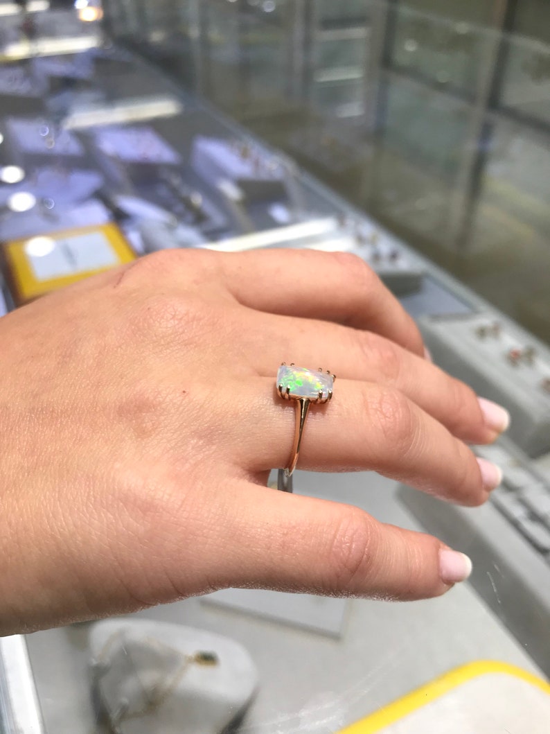 May include: A gold ring with a white opal gemstone. The opal is a rectangular shape with a slight curve. The ring is on a person's finger.