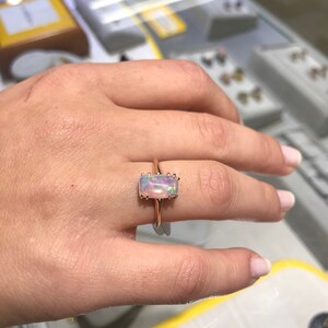 May include: A rose gold ring with a rectangular opal gemstone. The opal is iridescent with shades of pink, white, and blue.