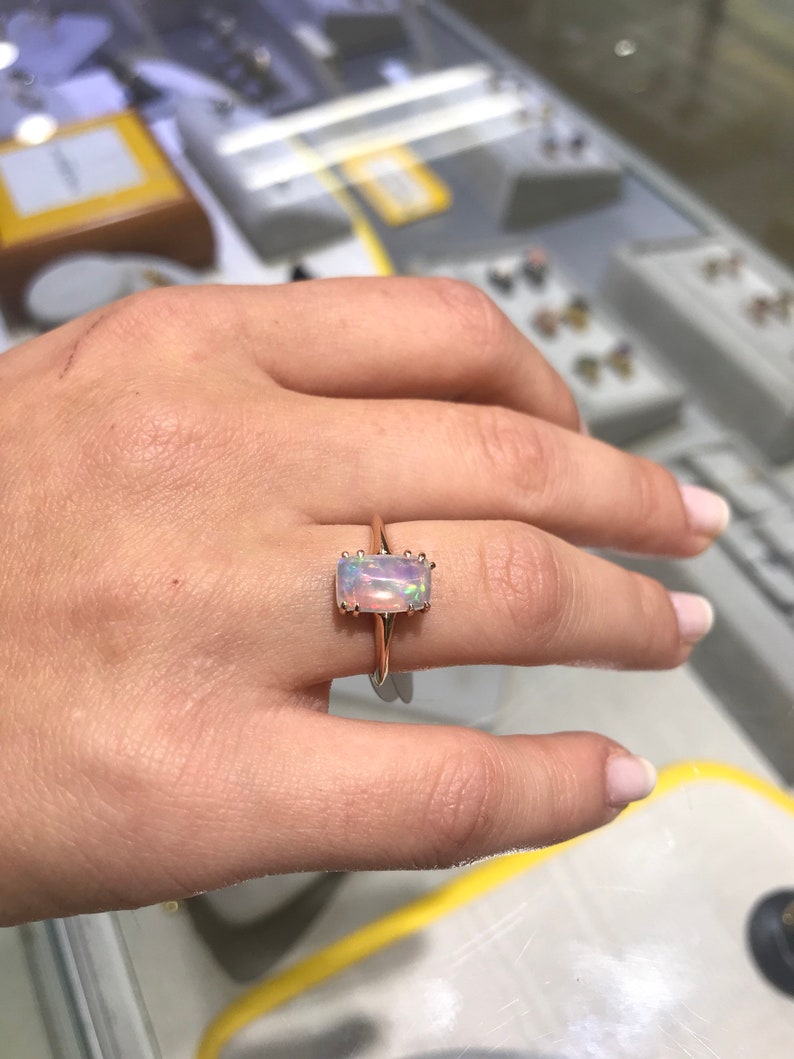 May include: A rose gold ring with a rectangular opal gemstone. The opal is iridescent with shades of pink, white, and blue.