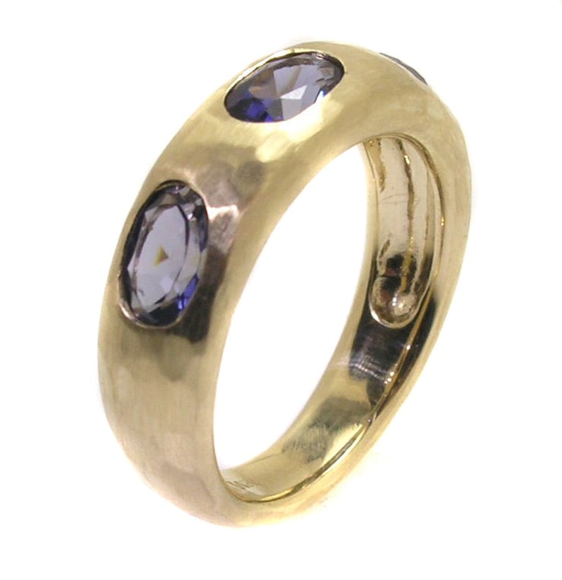 May include: A gold band ring with three purple gemstones set into the band. The ring has a hammered texture.