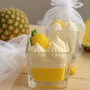 Pode incluir: Um recipiente de vidro quadrado preenchido com cera amarela, coberto com espirais de cera branca batida e um pequeno detalhe de cera em forma de abacaxi amarelo. A vela está parcialmente envolvida num saco de organza branco com um abacaxi.