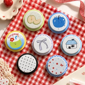 May include: Seven circular buttons with various designs including a green coconut with the text COCONUT, a light green apple, a blue apple, a purple bow, a house with a dog, black polka dots, and red stars on a blue and white checkered background.