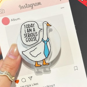 Puede incluir: Un soporte para teléfono de acrílico transparente con un dibujo de un ganso con una corbata azul y un bocadillo que dice "TODAY I AM A SERIOUS GOOSE". El soporte está sobre un fondo rosa con temática de Instagram.
