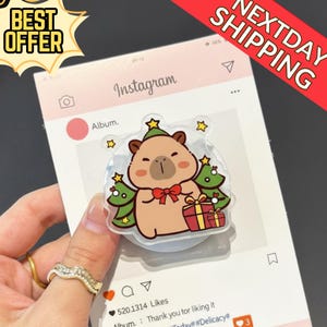 May include: A clear phone grip featuring a cartoon bear wearing a green hat and holding a gift. The bear is surrounded by green Christmas trees and yellow stars. The grip is on a pink Instagram-themed background with the text "BEST OFFER" and "NEXTDAY SHIPPING".