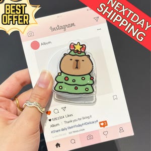 May include: A clear acrylic phone grip featuring a cartoon bear dressed as a Christmas tree. The grip is held against a pink Instagram-themed background with the text "BEST OFFER" and "NEXTDAY SHIPPING".