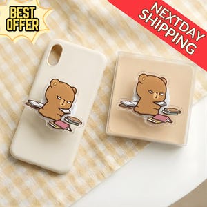 May include: A beige phone case with a brown bear-shaped phone grip. The bear is reading books. The image also includes a clear plastic case with the same bear grip and the text "BEST OFFER" and "NEXTDAY SHIPPING".
