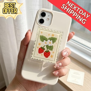 Può includere: Custodia per telefono color crema con un design a fragola su un adesivo trasparente. L'adesivo ha un design a francobollo con due fragole rosse, foglie verdi e fiori bianchi. È presente anche il testo "BEST OFFER".