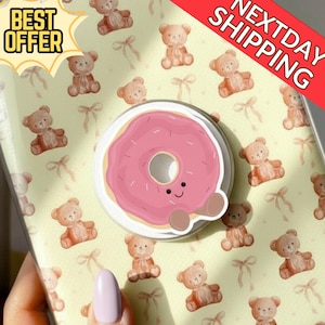Puede incluir: Un soporte para teléfono con un diseño de donut rosa con una cara sonriente. El soporte está adherido a una funda de teléfono con un patrón repetido de ositos de peluche y lazos. Las palabras "BEST OFFER" y "NEXTDAY SHIPPING" son visibles.