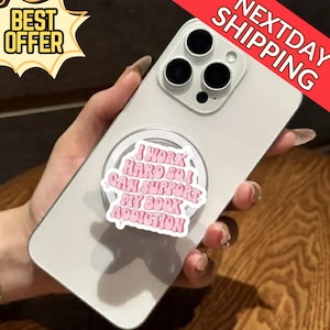 Puede incluir: Un smartphone gris claro con un soporte para teléfono que dice "I WORK HARD SO I CAN SUPPORT MY BOOK ADDICTION" en rosa y blanco. El teléfono tiene una cámara de triple lente. Se ve una pancarta de "BEST OFFER" y "NEXTDAY SHIPPING".