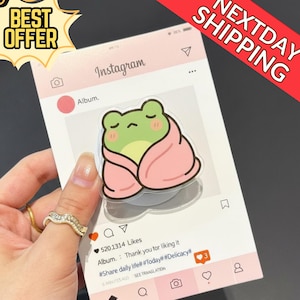 Może przedstawiać: Przezroczysty uchwyt do telefonu z kreskówkową żabą owiniętą w różowy koc, na różowym tle w stylu Instagrama. Tło zawiera logo Instagrama, ikonę serca i tekst "Album". Obraz zawiera również tekst "BEST OFFER" i "NEXTDAY SHIPPING."