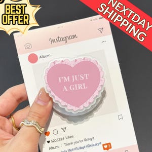 May include: A pink heart-shaped sticker with the text "I'M JUST A GIRL" is held up against a mock Instagram post. The sticker has a ruffled edge. The Instagram post is pink and white.
