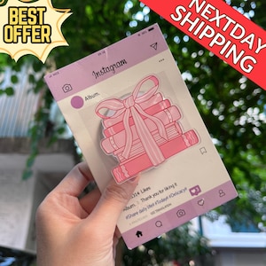 Könnte beinhalten: Ein rosa Aufkleber mit einem Stapel Bücher, die mit einer Schleife gebunden sind, im Stil eines Instagram-Posts. Der Aufkleber enthält den Text "Instagram" und "Album". Die Wörter "BEST OFFER" und "NEXTDAY SHIPPING" sind ebenfalls sichtbar.