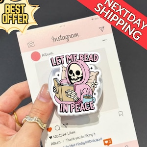 Könnte beinhalten: Ein klarer Handyhalter mit einem Cartoon-Skelett, das einen rosa Kapuzenpullover trägt, ein Buch liest und eine Tasse hält. Der Text "LET ME READ IN PEACE" wird angezeigt. Das Bild ist vor einem gefälschten Instagram-Post platziert.
