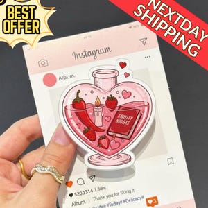 Peut inclure: Un autocollant en forme de cœur avec un design de potion rose, représentant un piment, des fraises, une bougie et un livre intitulé "Smutty Nights". L'autocollant est placé sur un fond rose sur le thème d'Instagram avec le texte "Best Offer" et "Next Day Shipping."