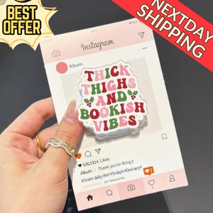 May include: A white and pink phone grip with the text "THICK THIGHS AND BOOKISH VIBES" in red, green, and pink. The grip is held in front of a pink Instagram-themed background. The image also includes the text "BEST OFFER" and "NEXTDAY SHIPPING".