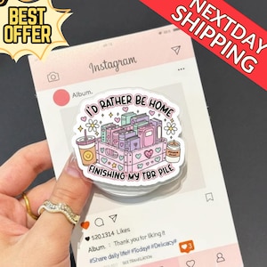 Puede incluir: Una pegatina transparente con el texto "I'D RATHER BE HOME FINISHING MY TBR PILE" con una pila de libros, una taza de café y una vela. La pegatina está sobre un fondo rosa con temática de Instagram.