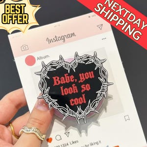 Puede incluir: Una pegatina negra en forma de corazón con el texto "Babe, you look so cool" en rojo, rodeada por un diseño de alambre de púas. La pegatina se muestra contra una interfaz de Instagram simulada.
